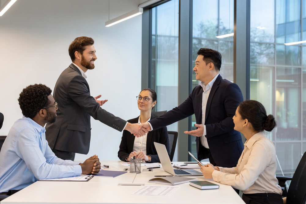 Two business leaders shaking hands across a conference table while colleagues look on